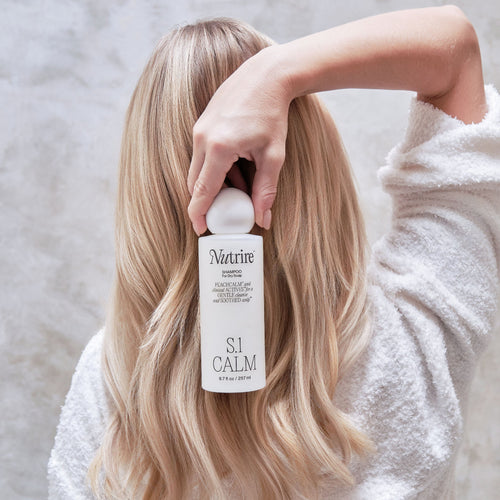 A woman with long blonde hair wearing a white bathrobe holds a bottle of Nutrine S1 Calm hair product in front of her face, set against a textured light gray wall.